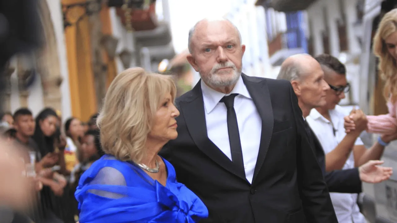 Mujer de cabello rubio con vestido azul camina del brazo de un hombre de traje negro.